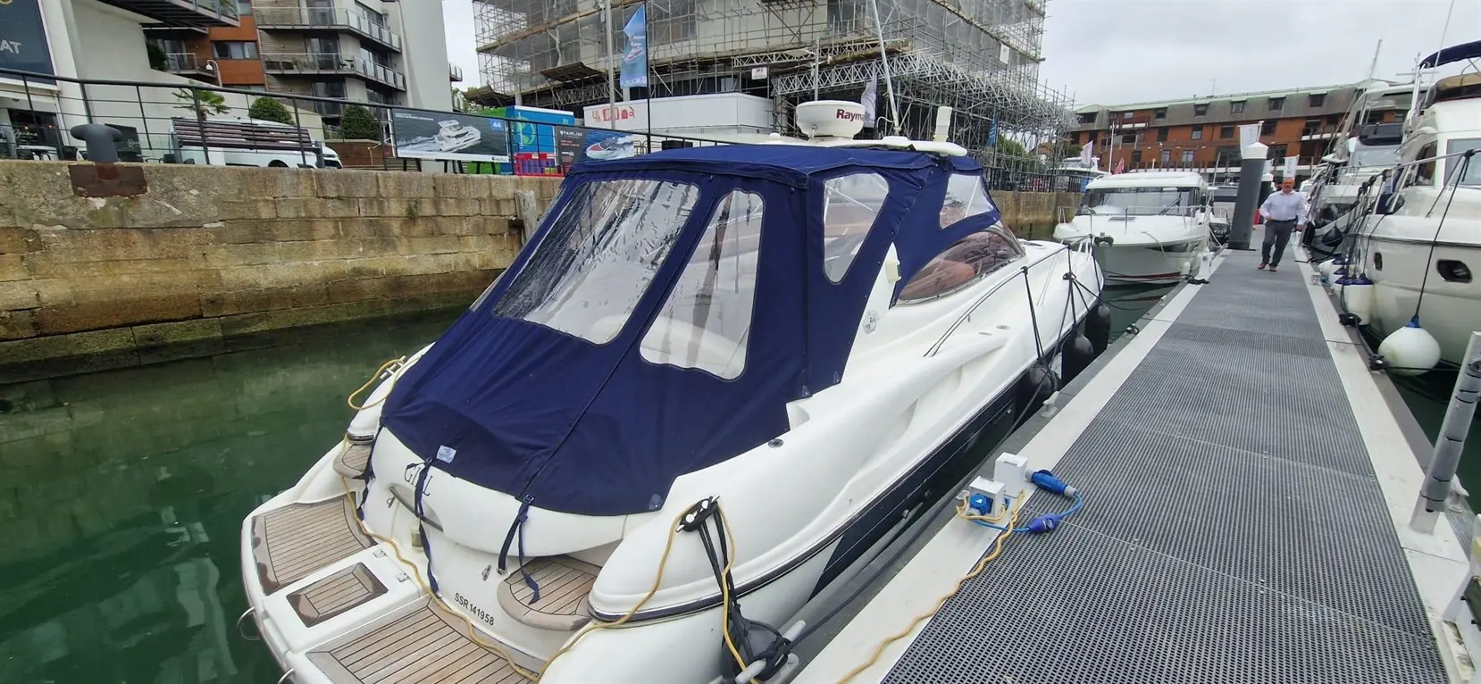A white motorboat with a blue canopy is docked at a marina, with a man walking nearby on the boardwalk.